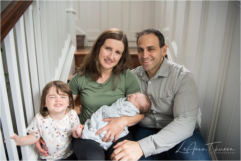 Parents sitting with their toddler daughter on the steps inside their beautiful home in Galloway, NJ holding their newborn son